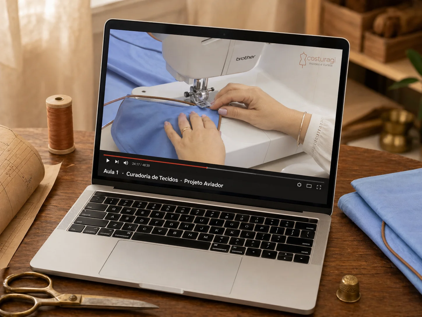 MacBook em bancada de ateliê mostrando Aula 1 - Curadoria de Tecidos do Projeto Aviador · logo Costuragi