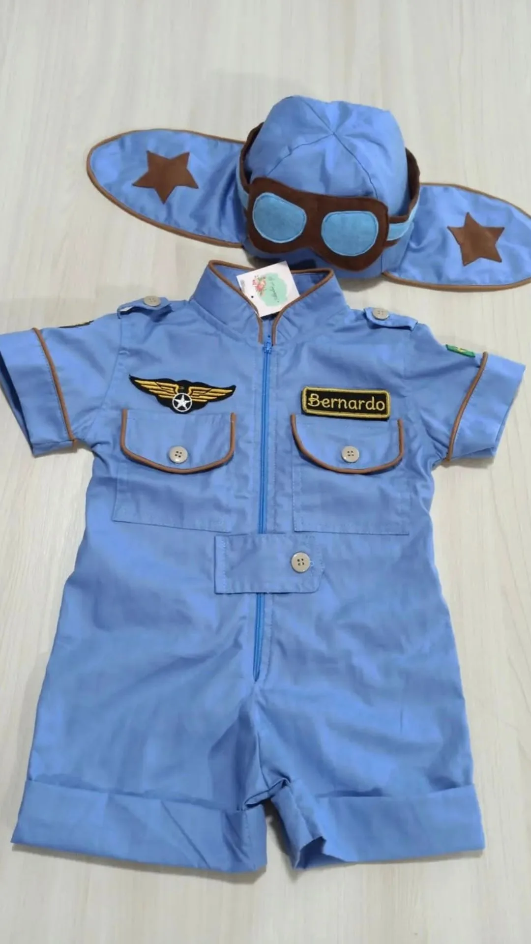 Macacão Aviador em sarja bege com touca combinando, viés contrastante cáqui, alça regulável com fivela metálica e bolso frontal com bordado de aviãozinho