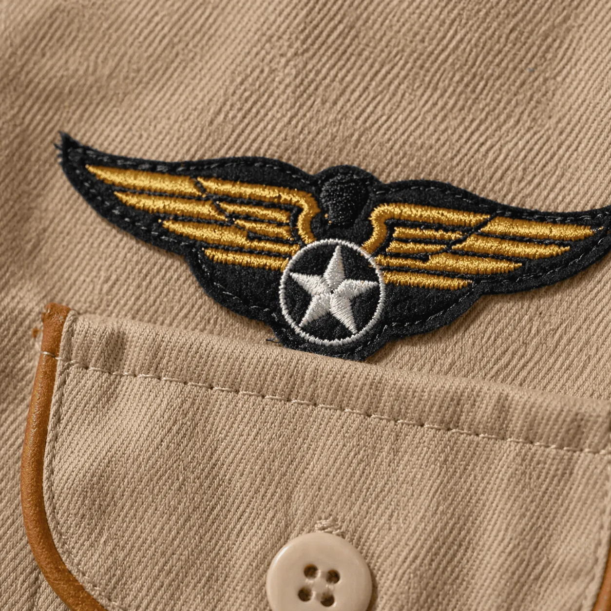 Macro do bordado de asas de aviador douradas com estrela branca central sobre o peito do macacão sarja bege, com bolso frontal e viés caramelo ao lado